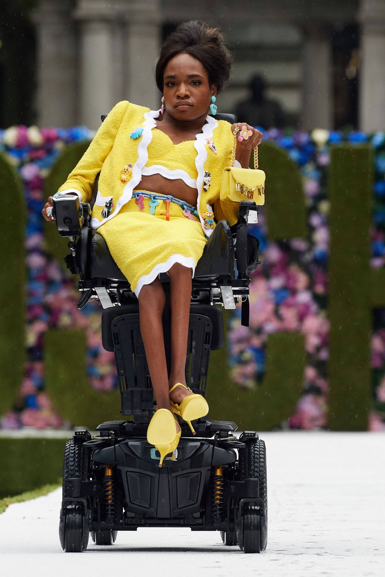 Woman in a wheelchair on a runway; she is wearing a canary yellow suit with a tube top, midi pencil skirt, and long sleeve jacket, each with a white scalloped edge. Her jacket is decorated with large stylized animal buttons, and her belt is made of colorful keys. Her open toed heels and small purse match her yellow suit. The purse is embellished with large gold charms spelling 'moschino' dangling from the purse flap. Her short black hair is parted in the center in front and teased up in the back.