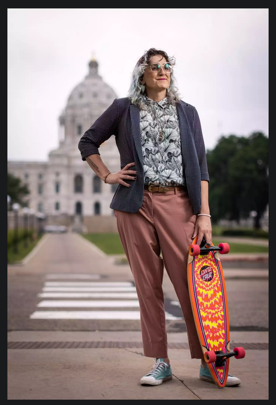 Woman standing with right hand on hip, left hand balancing a bright yellow and orange longboard against her knee. She is wearing her gray bobbed hair down in shoulder length curls. She is wearing a blazer with a miniscule pattern of blue and black checks over a button up shirt   covered in black and white stylized tropical leaves. Her mauve pants are held up with a thin brown western style belt and on her feet are sky blue chuck taylors.