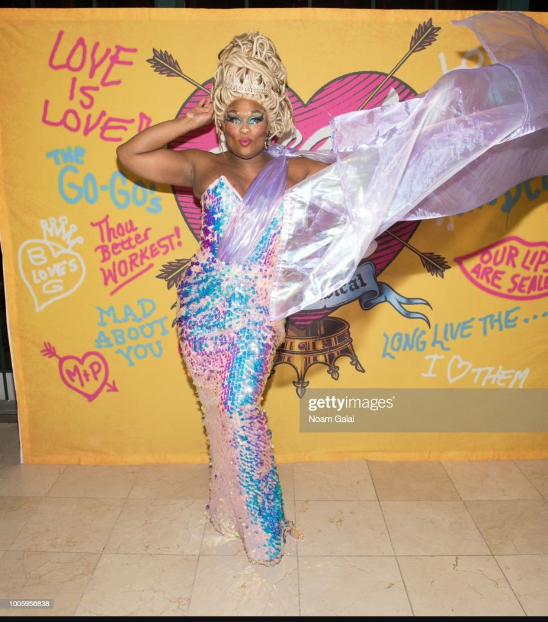 Woman wearing a floor length strapless glittering gown with a pointed bodice in shades of purple, pink, blue and magenta. Gathered iridescent lilac fabric extends from her waist, over her left shoulder, then cascades down her back into a train. She is standing with her left hand swooping her train in mid-air to the left, right hand on her head. Her hair is in blonde locs piled into a beehive with a tiara. Her blue eyeshadow and pink lips matches her dress.