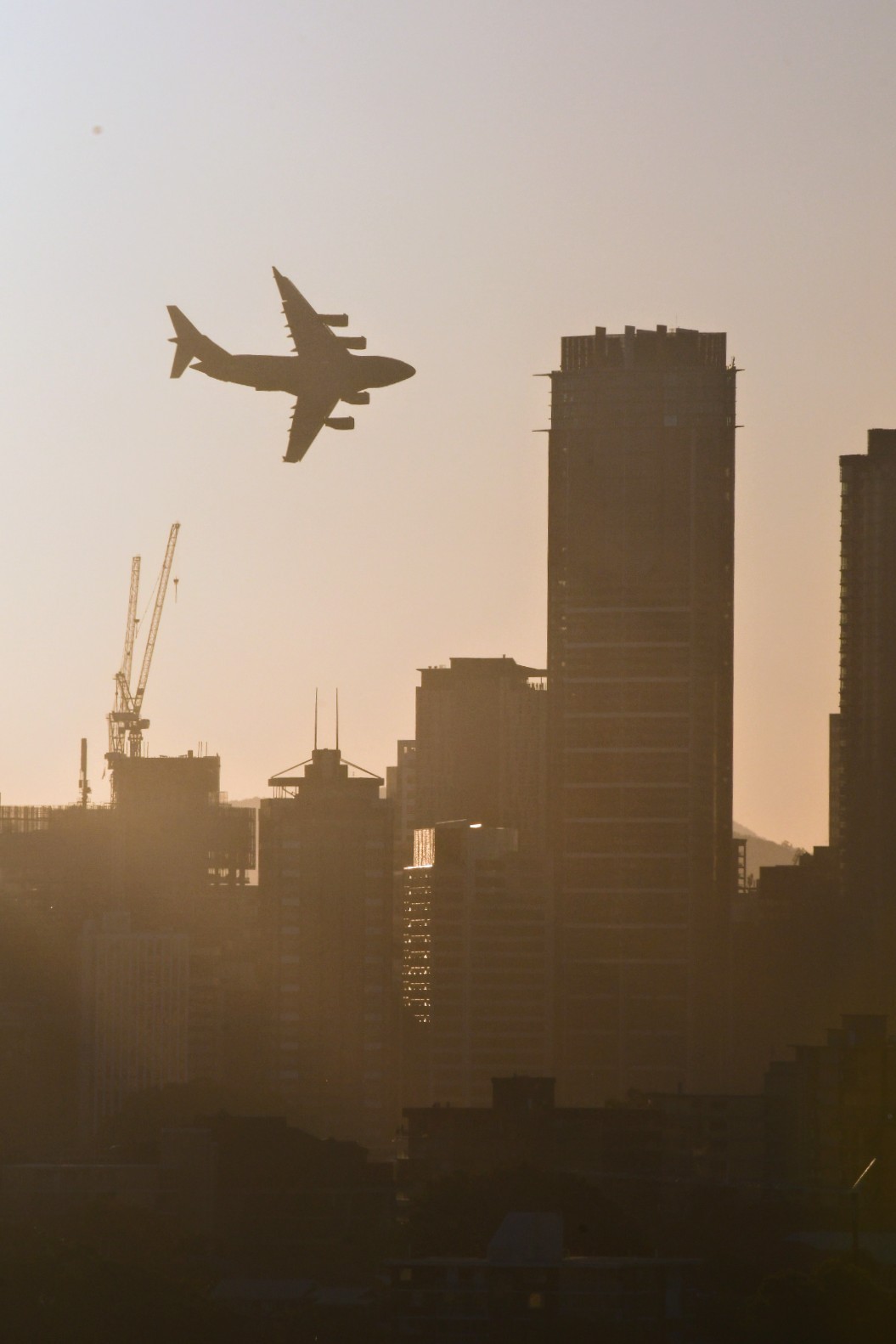 A photo of a large plane flying near tall city skyscrapers. The sky is tinted orange and the camera directed into the sun.